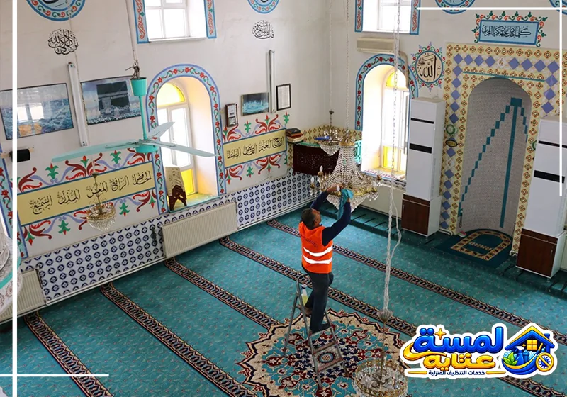 عامل ينظف ثريا المسجد باستخدام سلم للوصول إلى الأجزاء المرتفعة والعناية بالتفاصيل الدقيقة.